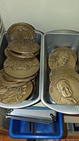 Photo showing two bins filled with stacked oversized bronze-toned commemorative coins and plaques with various portraits and designs.