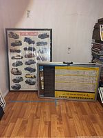 Two framed posters leaning against the wall on wooden floor; one prominently shows Rolls-Royce header and many classic car illustrations, the other shows a text-heavy poster.