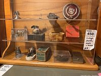 Three wooden shelves inside glass display, containing various vintage machine parts and automotive collectible emblems including a Cadillac emblem on top shelf.