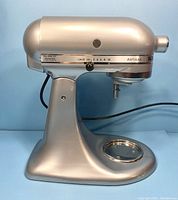 Front left side view showing speed control dial, power cord, tilt-head mechanism, and bowl mounting area, metallic chrome finish.