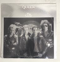 Front cover of The Game album by Queen. Shows four band members in black leather jackets on a silver reflective background.