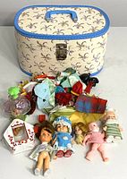 Shows vintage small dolls lined up with various doll clothes, accessories, and the decorated cream-colored oval carrying case with a blue floral pattern and top handle.