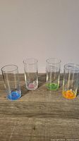 Four clear drinking glasses in a row showing different colored flower bottoms - blue, pink, green, orange.