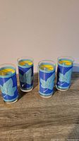 Four highball drinking glasses arranged side by side showing front side of leaf design with blue and light blue leaves on yellow background.