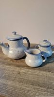 Blue speckled pottery teapot, sugar bowl, and creamer pitcher set on wooden surface, fully visible.