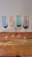 Front view of four colored champagne flutes placed on a wooden surface, showing light blue, amber, green, and smoky purple glasses from left to right.