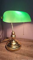 Full view of the vintage bankers style lamp highlighting the green glass shade and brass base with pull chain.