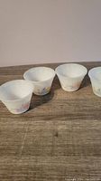 Four white milk glass small side bowls with pink, purple, and yellow floral patterns lined up on wooden surface.