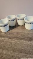 Four vintage Pyrex white coffee mugs with blue floral band design on the rim placed on a wooden surface.