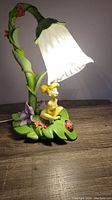 Lamp illuminated on wooden surface showing full lamp with leaf base, Tinkerbell figure, ladybug, vine stem, and tulip shade.