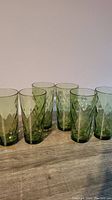 Six green highball glasses with diamond pattern arranged in two rows on a light wood surface against a plain pale wall.