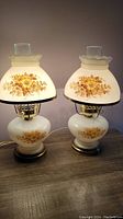 Pair of vintage milk glass lamps with floral painted designs on base and shade, placed side by side on table.