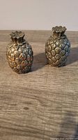 Pair of vintage metal pineapple-shaped salt and pepper shakers with textured surface and removable tops, photographed on a wooden surface.