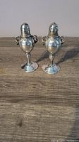 Front view of both silver plated salt and pepper shakers, showing embossed design, pedestal base, and perforated tops.