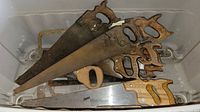 View showing a collection of handsaws stacked in a bin, handles mostly wooden, blades rusted and weathered.