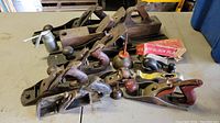 Top view of multiple antique block planers and hand planes arranged on a table with an oil can and a box labeled 'Auto Block Plane'. Visible wear and patina on tools.