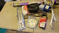 Photo shows various automotive and garden items on a table including a chrome bumper guard set, a garden tool with white handle, automotive belts and cables, a small black zippered case, and a silver dome shaped item.