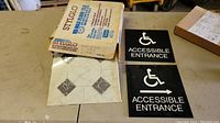Box of Stylo brand flooring tiles and two black accessible entrance signs with white wheelchair symbols and arrows, displayed on a table.