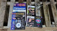 Top-down photo of music books and instructional DVDs with metal decorations and drum hardware parts arranged on wooden surface.