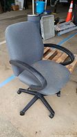 Gray office chair with curved black armrests and five-point caster base, visible dirt, placed indoors on concrete floor.