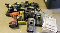 Photo showing nine power tools and accessories including four cordless drills, three battery chargers, one manual, and one smaller blue/silver tool. Items arranged on floor.