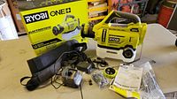 Ryobi ONE+ electrostatic sprayer with accessories spread on a table including charger, shoulder strap, nozzle parts, and manuals alongside original packaging box.