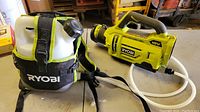 Photo showing the Ryobi electrostatic sprayer handheld unit and connected backpack tank with strap, placed on a surface.