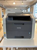 Front view of the two-tone grey Brother MFC-9130CW multifunction printer on a table, showing the scanner lid and paper output slot.