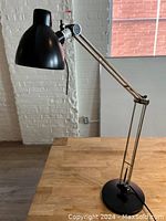 Black metal adjustable desk lamp on a wooden table with white brick wall background.