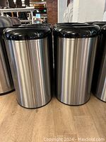 Two cylindrical stainless steel open-top garbage cans with black plastic lids on wooden floor