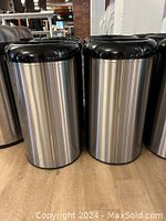 Two cylindrical stainless steel garbage cans with black plastic open tops on a wooden floor.