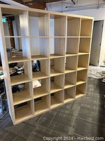 Light brown IKEA shelf unit with a grid of 25 cubes, placed indoors on gray carpet flooring next to other items.
