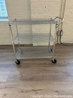 Full view of metal cart showing three wire shelves, handle, and wheels on hardwood floor against brick wall.