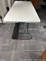 White folding table in unfolded position on carpeted floor, showing full length and metal legs beneath