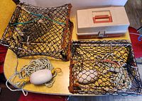 Photo showing two small crab traps with white floats and ropes next to an orange and white plastic tackle box on a table.