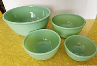Front angled view of four green Fire King Jadeite mixing bowls arranged on yellow surface, showing size and color of each bowl.