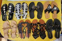 Top-down photo showing 8 pairs of women's sandals in two horizontal rows on a yellow surface, displaying various styles including gladiator, thong, and slip-on in black, white and tan colors