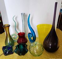 Wide view of nine colorful glass pieces of various shapes arranged on a yellow surface against a white background.