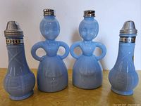 Four vintage salt and pepper shakers on a flat surface, including a pair of Merry Maids figurine-shaped shakers and two basketweave pattern shakers with visible labeling and tops.