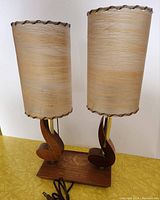 Front view of the pair of lamps showing cylindrical beige fiberglass shades with gold highlights and wooden scroll bases on a shared wooden platform.