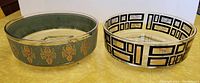 Two oval glass footed serving bowls with decorative rims, one teal with gold florals, the other white with black and gold geometric patterns, on a yellow surface.