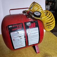 Full view of red KP Industries portable metal air tank showing two black instruction labels and attached yellow coiled air hose attached to top.