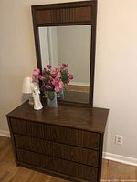Wooden bedroom bureau with three drawers and tall framed mirror against a light wall. Decorative items on top including flowers and a lamp.