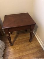 Solid wood bedroom side table with square top and turned spiral legs, shown in a corner on a wood floor.