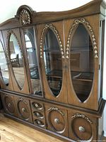 Front view of breakfront showing detailed oval glass doors with decorative gold-tone applique accents, wooden base cabinets and drawers, and ornamental top shell design.