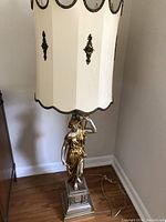 Full view of the lamp showing the sculpted woman base and cream-colored shade with gold-tone detailing, placed on wood floor against wall.