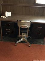 Photo shows dark metal desk with wood-like top surface, five drawers, and accompanying metal office chair with padded seat and backrest on caster wheels.