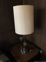 Metal table lamp with three clear globe bulbs and fabric shade on a wooden surface against paneled wall.