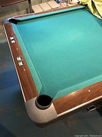 Pool table corner showing green felt playing surface, wood grain rails, and corner pocket