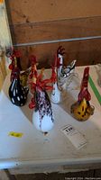 Group photo showing seven hand-blown art glass roosters in a variety of colors and patterns on a white surface with wooden backdrop
